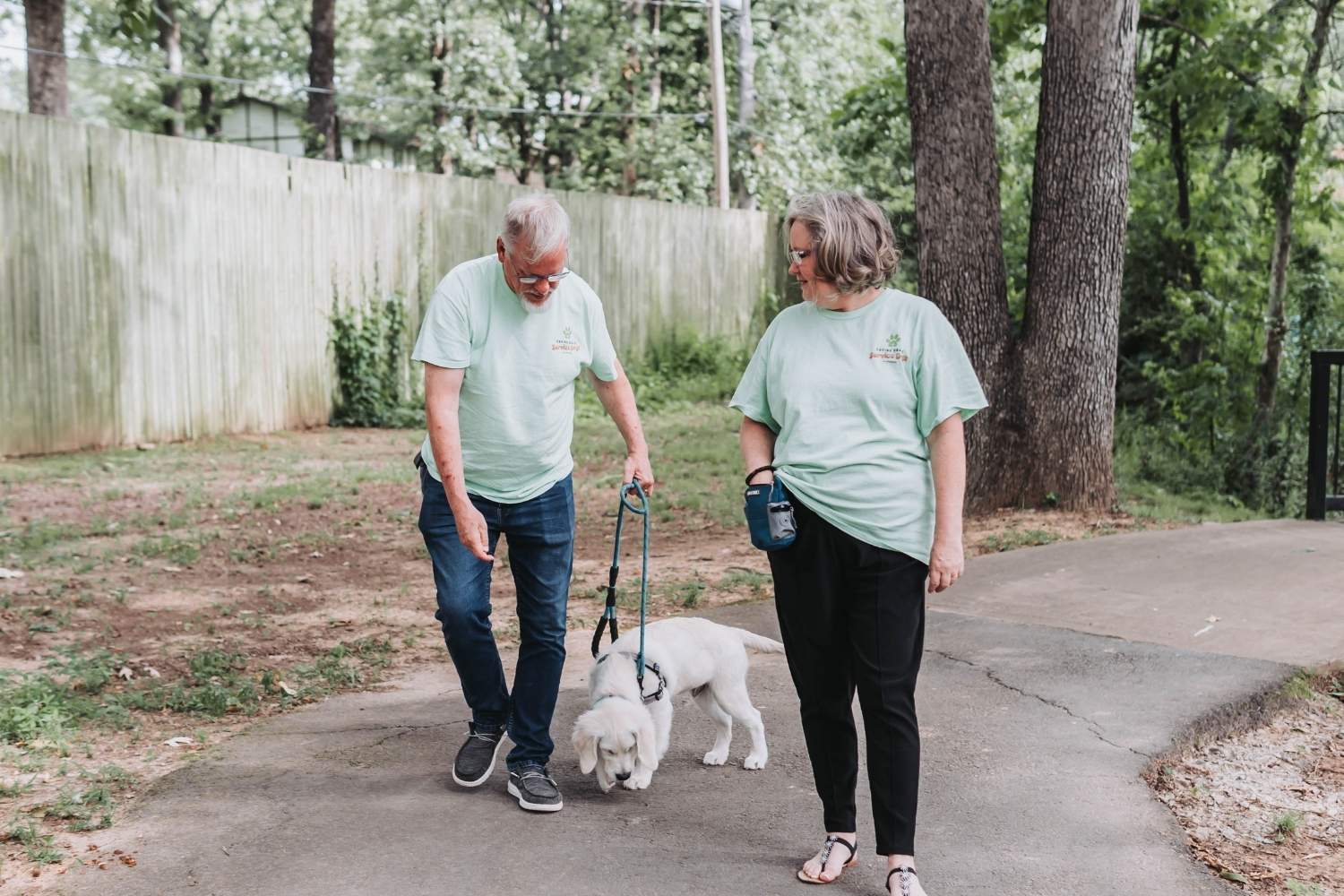 Two adults wearing matching light green shirts walk a white dog on a leash along a paved path in a wooded area. The man gestures toward the dog while the woman looks on.